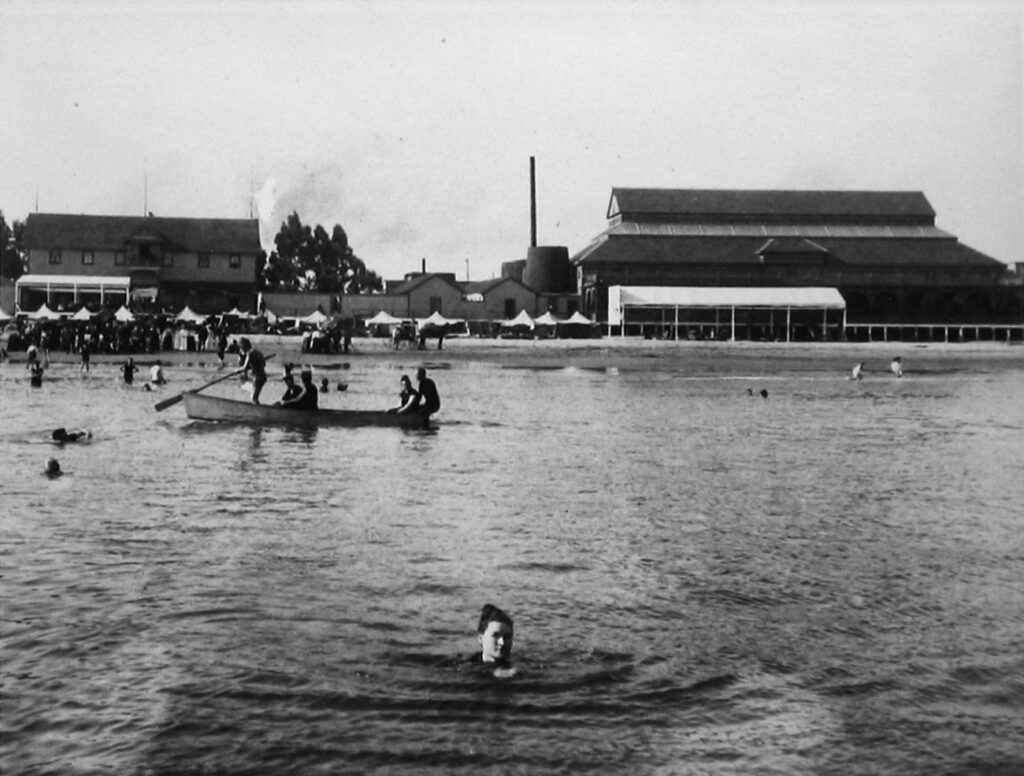 A woman swims far from the Santa Cruz shore in 1893, an unusual sight for the time. On the shore behind her is the Neptune Bath House (left) and the Miller-Leibbrandt Plunge Baths (right).